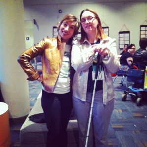 A woman in a gold jacket smiles next to a blind woman holding a cane wearing an oxford shirt and glasses. 